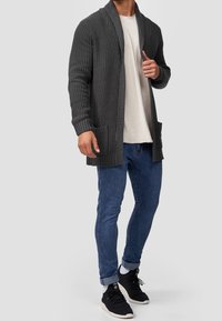 Hombre con un cárdigan de punto gris oscuro, camiseta blanca, jeans azules arremangados, calcetines blancos y zapatillas negras, de pie sobre un fondo claro.