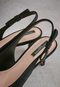 Mustat mokkanahkasta valmistetut slingback-korkokengät, joissa on terävät varpaat, pieni korko ja nauhalla varustettu rusetti. Kengissä on beige pohja, jossa on brändin logo.