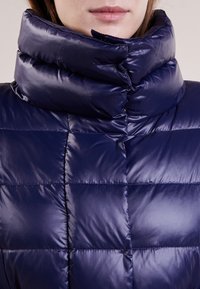 Marineblå pufferjakke med høj krave, quiltet design og skinnende tekstur. Materialet virker let og isolerende.