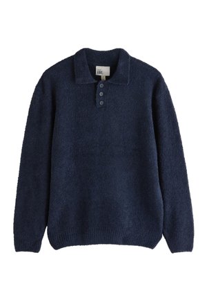 Ung mand står med armene krydset iført en navyblå sweater, lyseblå jeans og hvide sneakers mod en beige væg.