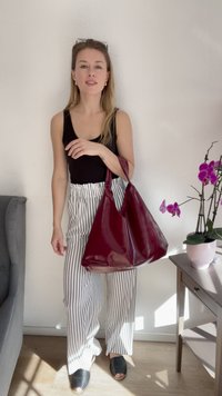 Burgundy patent leather tote bag with a wide opening and two shoulder straps; smooth texture with a glossy finish. Striped pants in the background.