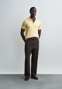 Muškarac stoji s rukama iza leđa, nosi svijetložutu polo majicu kratkih rukava, tamnobrede hlače i tamne kožne cipele na sivom podu.