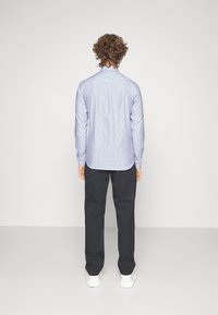 Camicia a maniche lunghe a righe azzurre con colletto dritto, abbinata a pantaloni scuri su misura e sneaker bianche, vista da dietro.