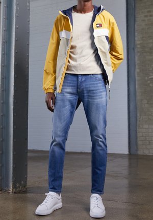 Farbenblockierte gelbe, weiße und marineblaue Jacke, hellbeiges T-Shirt, blaue Jeans und weiße Sneakers. Der Stoff wirkt leicht und lässig.