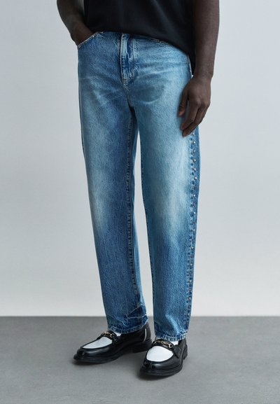 Homme portant un jean bleu avec des clous sur le côté et des mocassins noir et blanc, debout devant un fond gris uni.