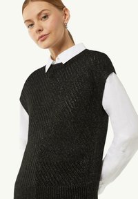 Czarny dzianinowy sweter bez rękawów o prążkowanej strukturze, akcentowany metalicznymi нитkami. Zestawiony z białą koszulą z kołnierzykiem pod spodem.