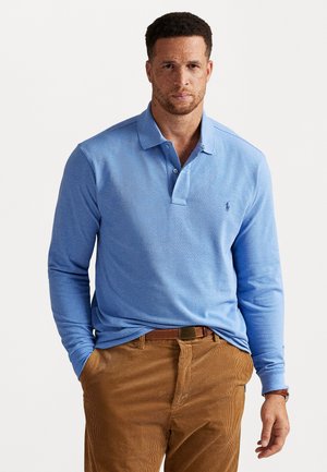 Lange mouwen, lichtblauwe polo-shirt van zachte stof, met een twee-knoopsluiting en klein logo op de linkerborst, gecombineerd met een paar bruine corduroypantalons.