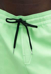 Ανοιχτό πράσινο μαγιό με μαύρο κορδόνι δεμένο σε φιόγκο στη ελαστική μέση, εμφανίζοντας λεπτομερείς ραφές.