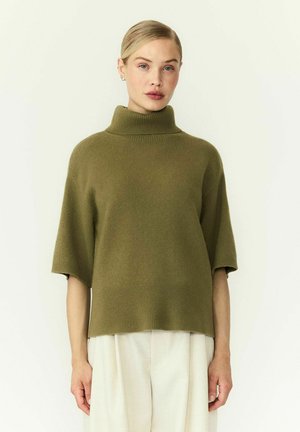 Femme aux cheveux blonds portant un pull à col roulé à manches courtes vert olive et un pantalon blanc ample, debout devant un fond clair uni.
