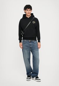 Felpa nera con logo, jeans dritti blu chiaro e sneaker bianco e nero, indossati con una piccola borsa a tracolla.