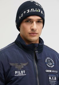 Navy knit beanie with white graphic text. The model wears a navy jacket featuring various embroidered patches and a zip closure.