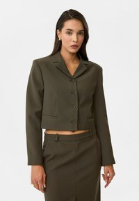 Blazer court vert olive avec un col à revers, six boutons sur le devant et poches latérales. Texture lisse, coupe ajustée et manches longues.