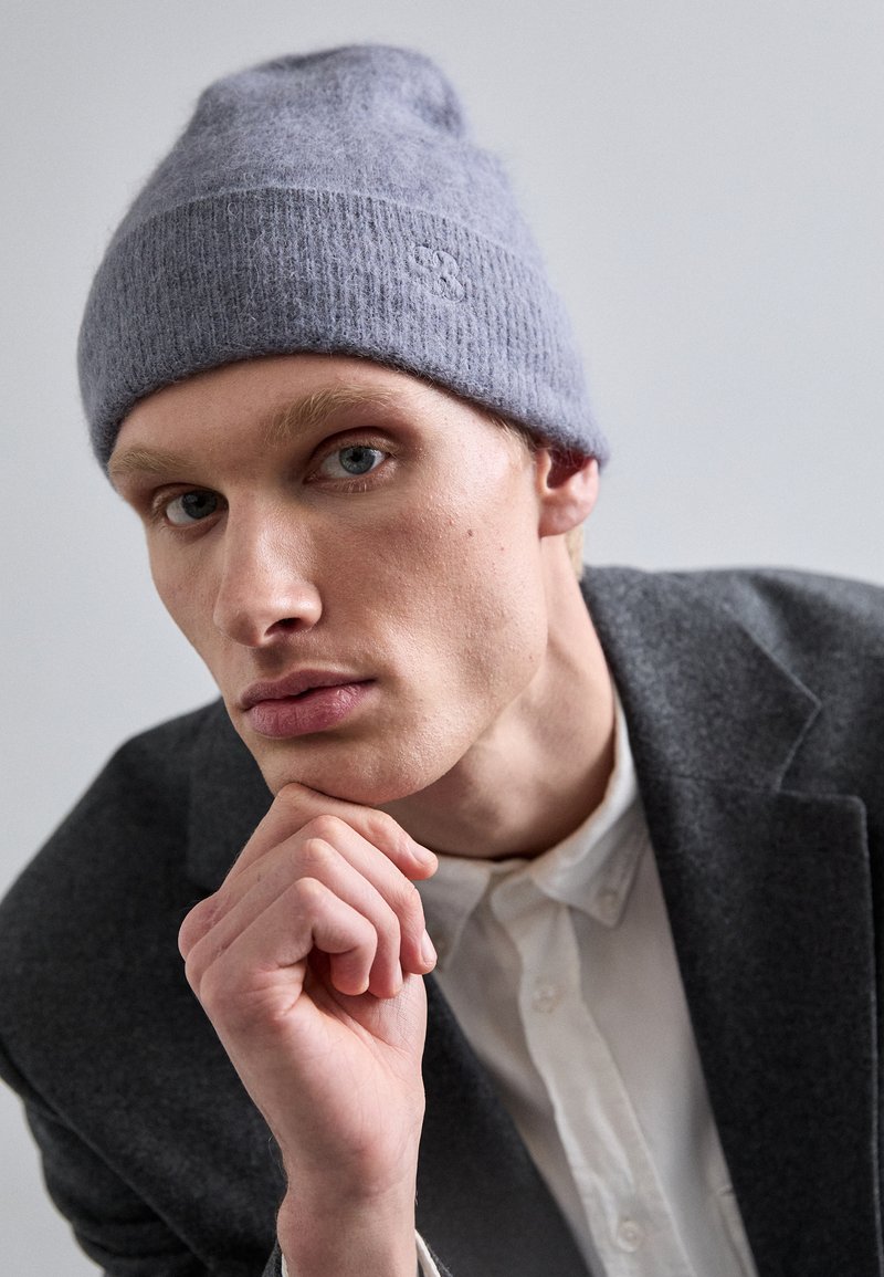 Beanie in lana grigia lavorata a maglia con texture a coste e dettaglio del logo, indossato leggermente scivolato. Lo sfondo presenta tonalità neutre.