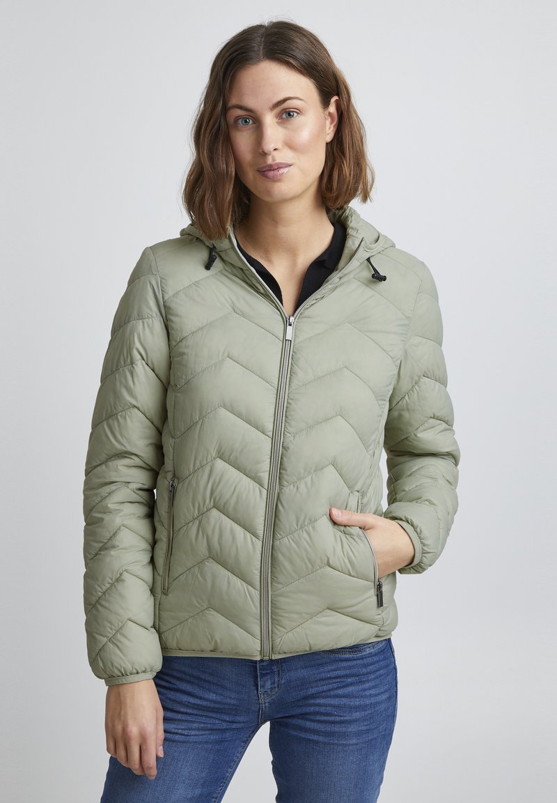 Veste matelassée verte claire avec un motif en chevron, fermeture éclair et capuche. Comprend deux poches latérales et des poignets élastiques. Portée par-dessus un haut noir.