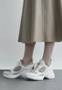 Bézs szoknyát viselő lábak fehér és arany hálós sportcipőben, vastag talppal, szürke padlón, világosszürke háttér előtt állva.