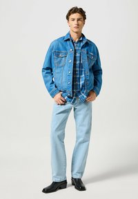 Veste en denim bleu clair avec deux poches poitrine et boutons cuivrés, superposée à une chemise à carreaux bleue. Jean bleu clair et bottes à talons noires.