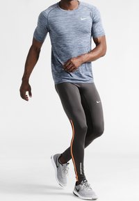 Herrarnas atletiska outfit som består av en blå texturerad kortärmad topp, svarta leggings med orangea sidopaneler och grå löparskor.