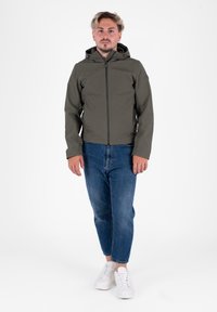 Giacca impermeabile verde oliva con cappuccio, chiusura con zip e due tasche laterali, abbinata a jeans blu slim-fit e sneakers bianche.