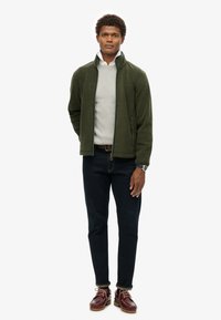 Giacca in pile verde con colletto alto, tessuto leggero, abbinata a un maglione grigio chiaro, jeans scuri e scarpe bordeaux con suola testurizzata.