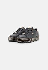 Sneaker in pelle nera con suola piatta spessa e a righe. Caratteristiche includono lacci neri, occhielli dorati e un accentuazione della parte posteriore in crema.