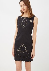 Joven mujer vistiendo un vestido negro sin mangas con intrincados patrones de encaje recortados en el pecho y el dobladillo, de pie frente a un fondo blanco liso.