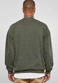 Hombre con cabello corto y tatuaje en el cuello, vistiendo sudadera grande de color verde oscuro, camiseta blanca debajo y pantalones beige, de espaldas frente a un fondo sencillo.