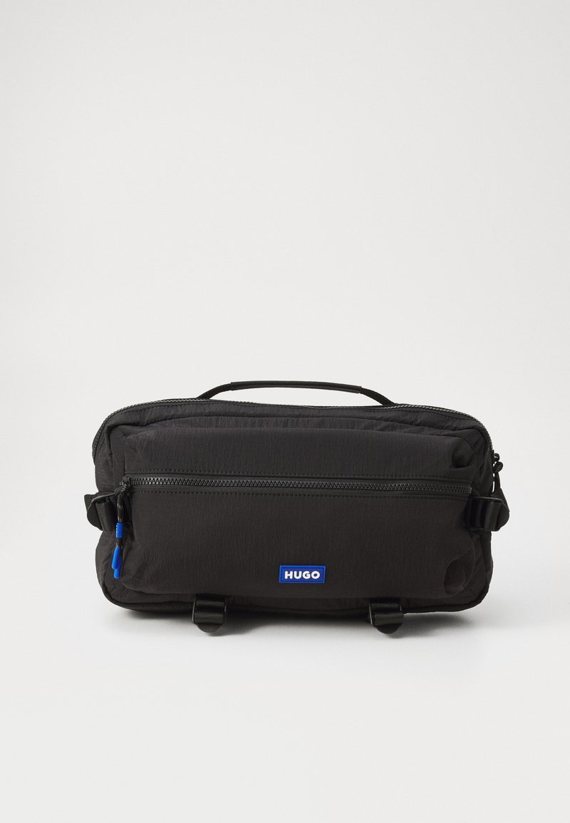 Sort nylon taske med en struktureret overflade, der har et håndtag i toppen, lynlåslukning, frontlomme og blå logoetiket. Kompakt og rektangulært design.