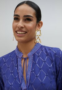 Kungblå klädesplagg med pärlade krage och diamantformade mönster. Guldörhängen med kvadratiska och triangulära mönster.