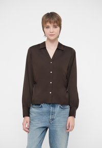 Noor särava lühikeste juustega noor inimene, kes kannab tumedast pruunist kangast nööbiga pikkade varrukatega särki ja helesiniseid teksaseid, seisab ühetoonilise tausta ees.