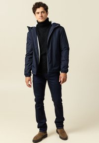 Giacca a vento blu navy con rivestimento in fleece bianco, con chiusura a zip e polsini elasticizzati, indossata sopra un dolcevita nero e jeans scuri. Stivali marroni.