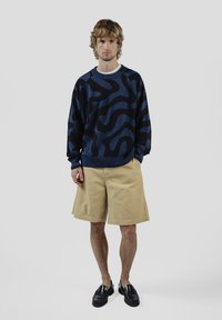 Laivastonsininen neule mustilla abstrakteilla kuvioilla, yhdistetty beigeihin shortseihin ja mustiin loafer-kenkiin. Neuleessa on pyöreä pääntie ja rento istuvuus.