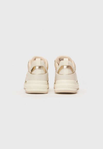 Zapatillas en crema y oro con una textura suave, forma redondeada, cuello acolchado y acentos metálicos en el talón y los laterales.