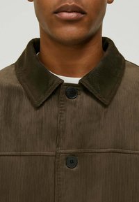 Giacca in corduroy marrone con collare a scollo a V, dotata di chiusura con pulsante nero e texture a coste verticali. Camicia bianca sottostante è parzialmente visibile.