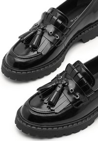 Μαύρα παπούτσια loafer από λουστρίνι με χοντρές λαστιχένιες σόλες, διακοσμητική φουντωτή λεπτομέρεια και διακοσμητικές φούντες μπροστά.