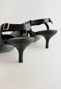 Schwarze Sandalen mit hohen Absätzen verfügen über ein glattes Lederoberteil, einen verstellbaren Knöchelriemen mit goldener Schnalle und ein spitz zulaufendes Absatzdesign.