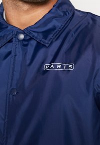 Navy blue waterproof jacket with a glossy finish, snap buttons, and a white "PARIS" logo stitched on the left chest area.