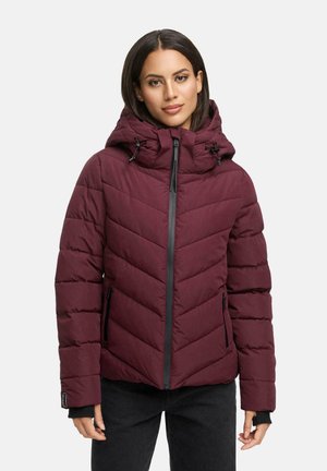 Vrouw die een bordeaux quilted pufferjack met een hoge kraag en capuchon draagt, helemaal ritself, gecombineerd met zwarte broek.