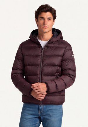 Giovane uomo che indossa una giacca imbottita trapuntata marrone scuro con cappuccio, in piedi con le mani giunte, vestito con jeans blu e camicia bianca sotto.