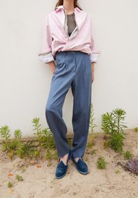 Osoba v volných modrých kalhotách, modrých sametových slip-on botách, s vrstvenými topy, stojí u bílé zdi se malými zelenými rostlinami poblíž.