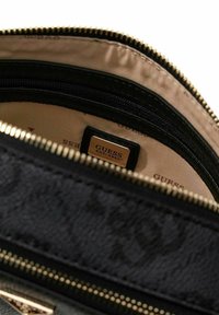 Portafoglio in tessuto nero con cerniera e dettagli dorati, con interno beige e un'etichetta in pelle nera con il marchio "GUESS."