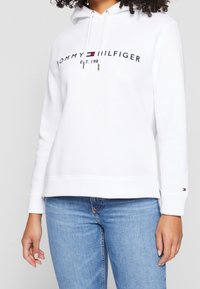 Felpa bianca realizzata in materiale morbido, con una grafica frontale che presenta "TOMMY HILFIGER" e il logo. Include un cappuccio regolabile con lacci e polsini a costine.