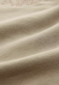 Beige bomuldsstof med en glat tekstur, med subtile folder og et trykt design i lysebrun, der giver et blødt og let udseende.