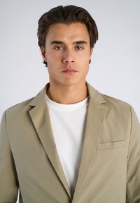 Νεαρός άνδρας με κοντά σκούρα μαλλιά φοράει ένα μπεζ σακάκι πάνω από ένα λευκό μπλουζάκι με στρογγυλή λαιμόκοψη, κοιτάζοντας μπροστά σε απλό ανοιχτό φόντο.