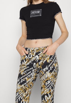 Vrouw draagt een zwart Versace Jeans Couture cropped T-shirt en zwarte broek met gouden barokpatronen en witte logo-tekst, handen in de zakken.