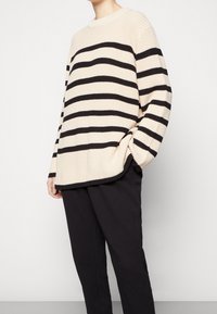 Kremowo-czarny sweter w paski z dzianiny z okrągłym dekoltem, luźnym krojem i ściągaczami na rękawach. Nosi się go na czarne spodnie z fakturą.