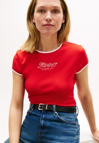 Κόκκινο κοντομάνικο T-shirt με λευκή επένδυση, με το λογότυπο "TOMMY HILFIGER". Συνδυασμένο με ψηλόμεσο μπλε τζιν και μαύρη ζώνη.