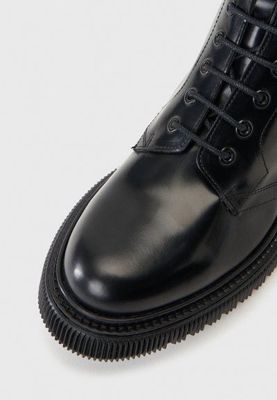 Κοντινό πλάνο ενός γυαλισμένου μαύρου δερμάτινου μποτάκι με κορδόνια και χοντρή, εξογκωμένη λαστιχένια σόλα σε ανοιχτό φόντο.