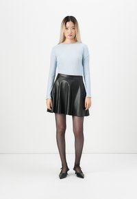 Žena s dlouhými blond vlasy, oblečená v světle modrém strukturovaném topu s dlouhými rukávy, černé kožené sukni, průhledných černých punčochách a černých špičatých botách.