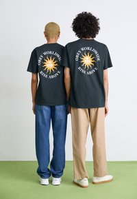 Svarte grafiske t-skjorter med soldesign, med teksten "Obey Worldwide" og "Rise Above". Båret med avslappede blå jeans og beige bukser.