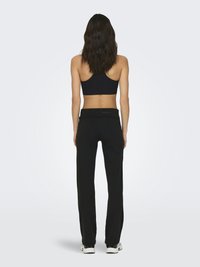 Femme aux longs cheveux foncés portant un soutien-gorge de sport noir, un pantalon noir et des baskets blanches, debout de dos sur un fond clair uni.